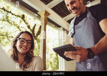 Junge Frau in einem Café, die über das Menü bestellt, während der Kellner mit einem digitalen Tablet bestellt. Lächelnder Kellner in der Schürze, der am Tisch steht und weitere Informationen hinzufügt Stockfoto