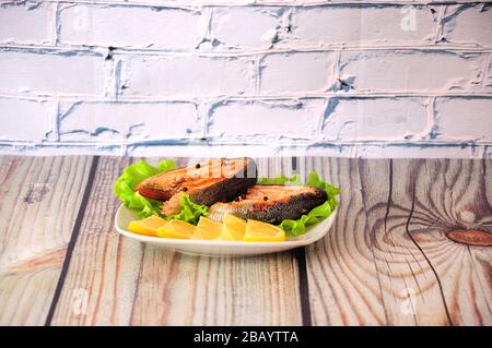 Zwei gegrillte rosafarbene Lachssteaks liegen auf einer weißen Keramikplatte, die mit Zitronenscheiben und einem Salatblatt verziert ist, die ganze Komposition auf einem Holztisch. C Stockfoto