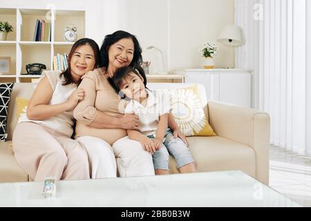 Fröhliche vietnamesische seriöse Frau mit ihrer erwachsenen Tochter und ihrer kleinen Enkelin, die zu Hause auf dem Sofa sitzt Stockfoto