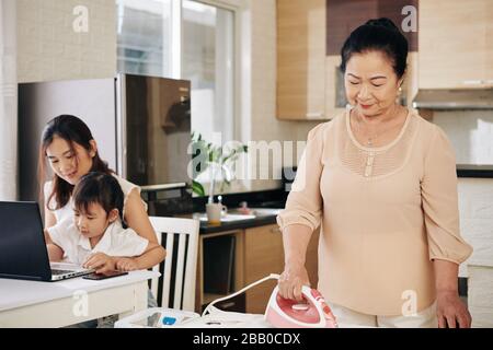 Junge Asianerin, die mit ihrer Tochter am Computer arbeitet, während ihre ältere Mutter ihre Kleidung bügelt Stockfoto