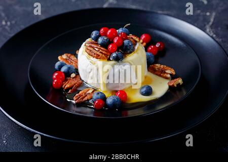 Partystarter: Gebackener brie-käse mit Pecan, Blaubeeren, Preiselbeeren und Ahorn-Sirup auf einem schwarzen Teller auf einem dunklen Betontisch, Stockfoto