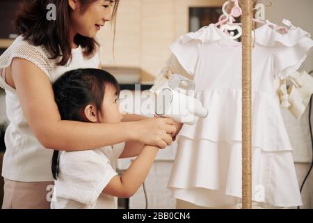Die junge Asianerin, die ihrer kleinen Tochter beigebracht hat, wie man Kleidung dampft Stockfoto