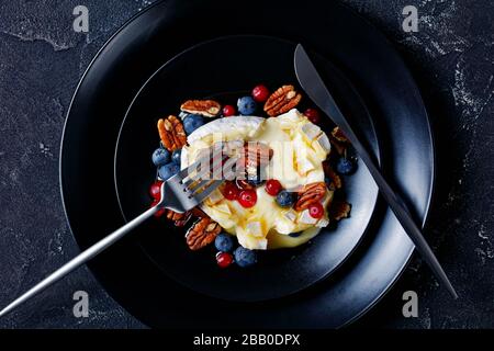 Ahorn Pecan gebackene brie mit Beeren: Blaubeeren, Preiselbeeren und Honig auf einem schwarzen Teller mit silberfarbenem Besteck auf einem dunklen Betonbackgroschen Stockfoto