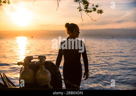 Weiblicher Tauchlehrer für Scuba, der einen nassen Anzug trägt, der neben einem Zwillingstank bei Sonnenuntergang steht Stockfoto