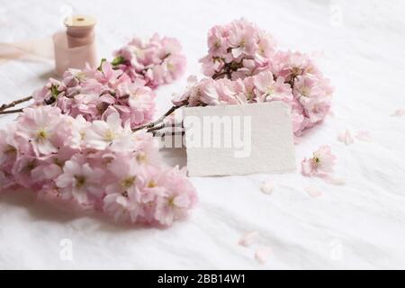 Hochzeitstafel-Szene. Leeres Grußgeschäft, RSVP-Baumwollpapierkarte auf Leinentisch Hintergrund mit rosa blühendem Kirschbaum Bran Stockfoto