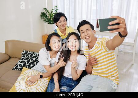 Fröhlicher, gutaussehender junger Mann, der selfie mit seinen Freunden nimmt und dabei Fingerherzgeste macht, wenn sie zu Hause auf einem großen Sofa sitzen Stockfoto