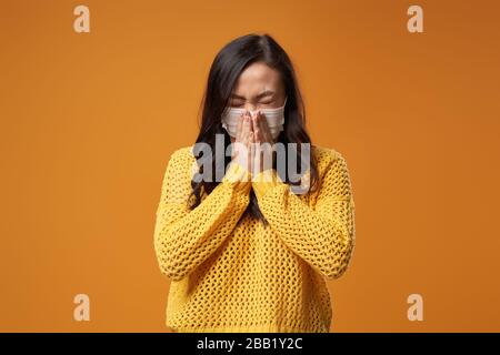 Maskierte brünette Frau niest, während sie im Studio auf leerem orangefarbenem Hintergrund steht. Stockfoto