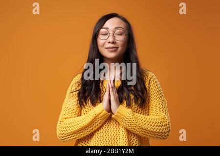 Betende junge asiatin mit geschlossenen Augen, die eine Brille trägt, und in gelbem Strickpullover auf leerem orangefarbenem Hintergrund im Studio Stockfoto