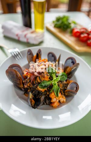 Fisch und Meeresfrüchte Squid Ink Pasta mit Muscheln Garnelen und Tintenfisch auf weißem Teller Tomatenolivöl und Essig im Backgroog auf einem grünen Holztt Stockfoto