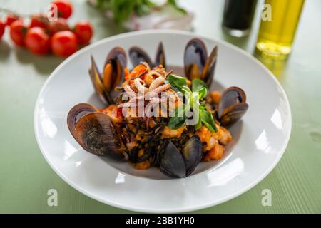 Fisch und Meeresfrüchte Squid Ink Pasta mit Muscheln Garnelen und Tintenfisch auf weißem Teller Tomatenolivöl und Essig im Backgroog auf einem grünen Holztt Stockfoto