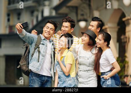 Fröhlicher vietnamesischer junger Mann, der selfie mit einer Gruppe von Freunden nimmt, wenn er auf der Straße steht Stockfoto