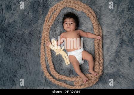 Ganzkörperbild des Jungen, der auf einem grauen Pelz liegt und ein Kaninchenspielzeug hat, neugierig auf den Zuschauer schauend. Stockfoto