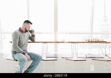 Junger bärtiger Mann im Café. Er kleidete sich mit grauem Pullover und blauer Jeans. Sitzt auf einem Hochstuhl am langen Tisch in der Nähe des Fensters. Mann trinkt Getränk mit Grün Stockfoto