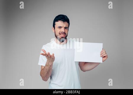 Junger Mann isoliert über Hintergrund. Guy zögert mit der Kamera. Emotionaler Ausdruck im Studio. Halten Sie die weiße leere Seite in den Händen. Brünette fühlen sich verkleidet. Stockfoto