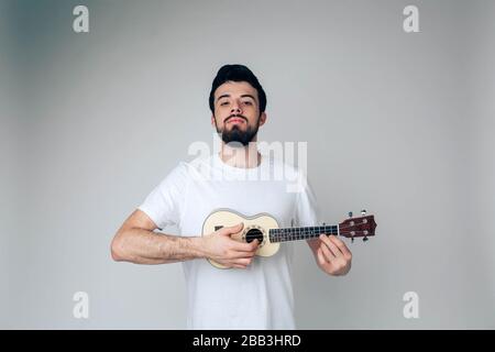 Junger Mann isoliert über Hintergrund. Lustiger seriöser Kerl mit Ukulele in den Händen. Ruhiger, ruhiger, stolzer Musiker, der posiert Stockfoto