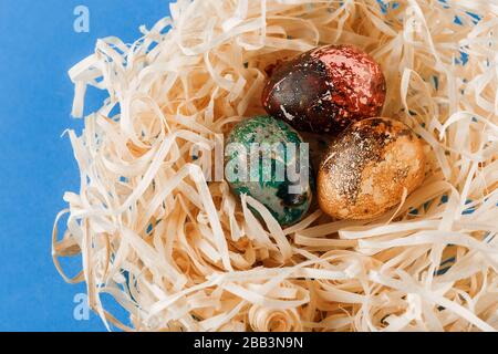 Drei ostereier sind rot, gelb und grün lackiert. Eier liegen in einem Holzspäne. Bemalte ostereier auf blauem Grund. Nahaufnahme. Stockfoto