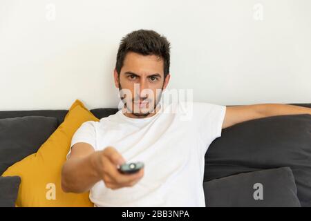 Ein Mann sitzt zu Hause auf der Couch und beobachtet Fernsehen. Er hält eine Fernbedienung und versucht, einen interessanten Kanal zu finden. Stockfoto