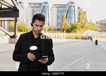 Ein junger gutaussehender Mann steht in einem schwarzen Business-Anzug auf dem Hintergrund eines Bürogebäudes. Er trinkt seinen Kaffee aus einer nachfüllbaren Tasse und nimmt Stockfoto