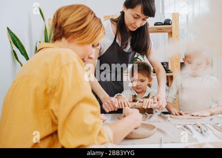 Familien-Meisterklasse in der Töpferei. Im Studio skulptieren zwei Kinder und zwei Frauen. Blick durch eine getrocknete braune Pflanze Stockfoto