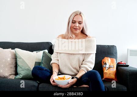 Eine einzelne blonde kaukasische Frau, die auf einem Sofa sitzt und eine weiße Schüssel mit tangiertem Käse Doritos hat Stockfoto