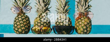 Nahaufnahme einer Gruppe frischer Ananas-Hüftfamilie in lustiger und stilvoller Sonnenbrille auf dem Tisch vor blauem hellem Holzhintergrund. Funky, reif, modisch Stockfoto