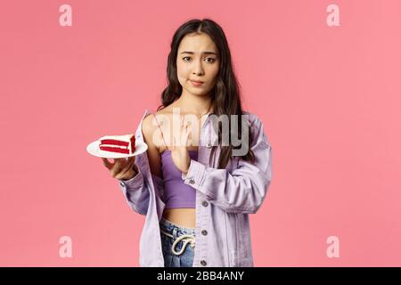 Portrait des skeptischen jungen asiatischen eleganten schlanken Mädchens, das sich weigert, Kuchen zu essen, Smirk zeigen Top-Zeichen zum Dessert auf dem Teller liegen und versuchen es ablehnen, enttäuscht Stockfoto