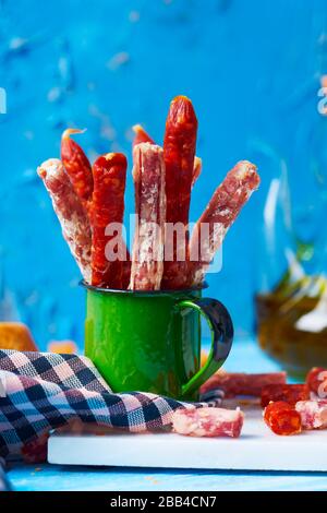 Einige Fuet- und Chorizo-Stücke, zwei verschiedene spanische Wurstwaren in einem grünen Becher auf einem Tisch, neben einigen Brotscheiben und einem Grausamkeiten mit Olive o Stockfoto