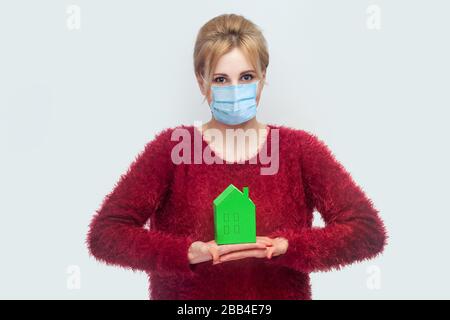 Bleiben Sie zu Hause, bleiben Sie sicher. Quarantänezeit. Porträt der jungen Frau mit chirurgischer medizinischer Maske in roter Bluse stehend, Papier halten kleines Haus. Innenbereich Stockfoto