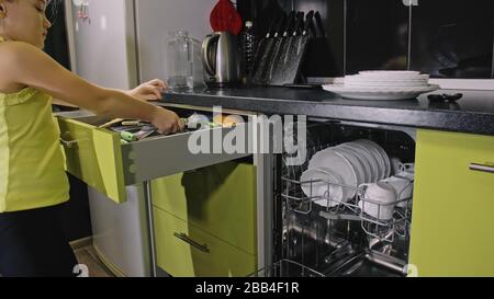 Schlaue Mädchen lernen, Geschirrspüler zu benutzen. Stilvolle, moderne Einbauküche in grünem Schwarz. Kind legt sauberes Geschirr. Stockfoto