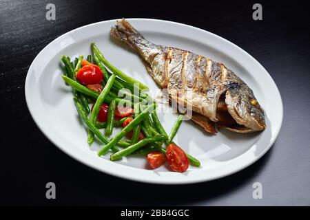 Von oben Blick auf gebackenen Seevoss mit frischem Salat grüner Nadelbohnen und Kirschtomaten auf weißem Teller. Leckeres Gericht auf schwarzem Tisch. Konzept der Restaurantküche, Meeresfrüchte. Stockfoto