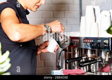 Ein Barist in einer schwarzen Schürze gießt frisch zubereiteten Kaffee vor dem Hintergrund einer Kaffeemaschine und einer Rutsche leerer Papierbecher in eine Papierbecher. Stockfoto