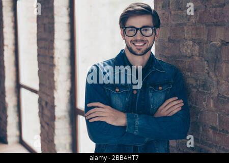Erfolgreicher bärtiger Student im legeren Jeans-Outfit steht in der Nähe der Fenster- und Ziegelwand, trägt eine Brille und lächelt mit gekreuzten Händen, innen Stockfoto
