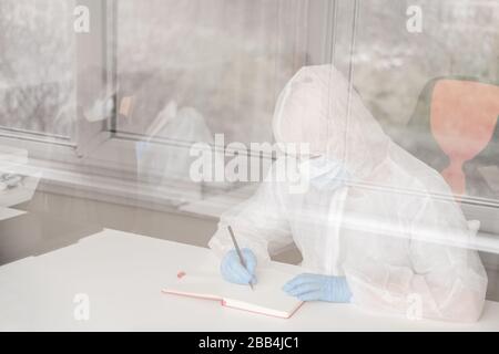 Mädchen im schützenden weißen, lichtdurchlässigen Anzug, blaue Gummihandschuhe, medizinische Maske sitzt zu Hause am Tisch und zeichnet im Skizzenbuch mit Bleistift. Remote-Arbeit von Stockfoto