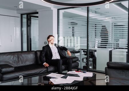 Eine neue Idee zu entwickeln... ein gutaussehender und erfolgreicher Geschäftsmann im eleganten Anzug denkt an etwas, während er im Büro auf dem Sofa sitzt Stockfoto