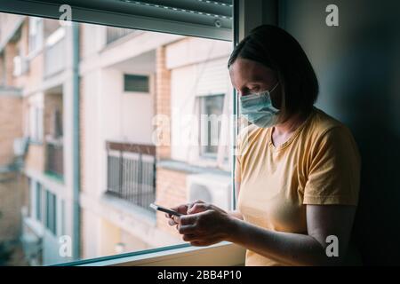 Frau in Selbstisolation bei Virusausbruch mit Handy. Besorgte weibliche Person mit chirurgischer Schutzmaske, Smartphone halten, selektiv f. Stockfoto