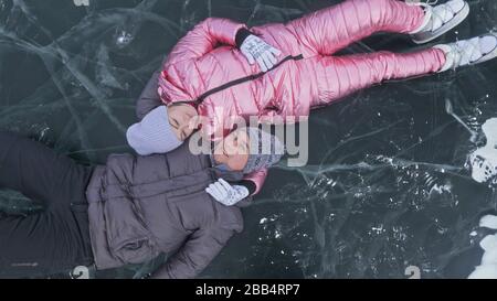 Das Paar hat Spaß beim Winterspaziergang vor dem Eis des zugefrorenen Sees. Liebhaber liegen auf klarem Eis, haben Spaß, küssen und umarmen sich. Ansicht von oben. Stockfoto