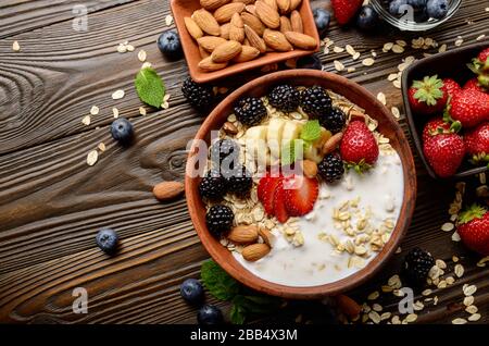Flach top Blick auf Obst gesund Müsli mit Banane Erdbeere Mandeln und Blackberry mit Joghurt in Ton Teller auf hölzernen Küchentisch Stockfoto