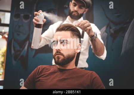 Friseur sprüht Lack auf die Haare des Kunden. Friseur macht Styling ein junger Hipster Stockfoto