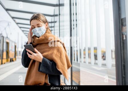 Junge und depressive Frauen in Gesichtsmaske sorgten sich, dass sie während einer Epidemie schlechte Nachrichten auf einem Smartphone an einem Stopp der öffentlichen Verkehrsmittel lesen. Konzept der Panik und des schlechten Einflusses der Nachrichten Stockfoto