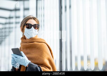 Junge Frau in Gesichtsmaske, medizinische Handschuhe und Sonnenbrille mit einem Smartphone an der Haltestelle der öffentlichen Verkehrsmittel während einer Epidemie. Konzept der sozialen Distanz und Online-Kommunikation Stockfoto