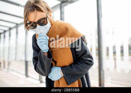 Frau in Gesichtsmaske fühlt sich krank an und hustet an einem öffentlichen Verkehrshalt im Freien. Konzept der Coronavirus Epidemie Stockfoto