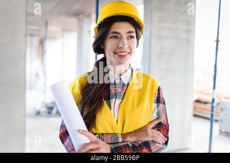 Positives Lächeln Frau Ingenieur Baumeister steht auf einer Baustelle mit Papier mit Zeichnungen Stockfoto