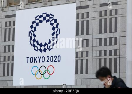 Tokio, Japan. März 2020. Ein Mann, der Schutzmasken trägt, geht am Logo der Olympischen Spiele in Tokio 2020 vorbei, das an einer Wand des Tokyo Metropolitan Government Building in Tokio, Japan, am 25. März 2020 gezeigt wird. Die Olympischen Spiele in Tokio wurden auf den 23. Juli bis 8. August 2021 verschoben, die Organisatoren der Olympischen Spiele 2020 in Tokio und der Paralympischen Spiele wurden am Montag bekannt gegeben. Credit: Du Xiaoyi/Xinhua/Alamy Live News Stockfoto