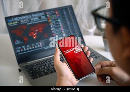 Coronavirus oder Covid-19 Pandemie News Update Hintergrundkonzept. Verspotzen Sie Ihr Mobiltelefon, eine Person, die Smartphoe hält und auf den Bildschirm des Laptops starrt Stockfoto