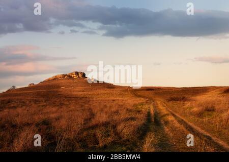 Getrocknete Felder auf dem Hintergrund eines kuriosen steinigen Berges unter dem übergiebelten Himmel. Das Foto wird zur goldenen Stunde aufgenommen. Stockfoto