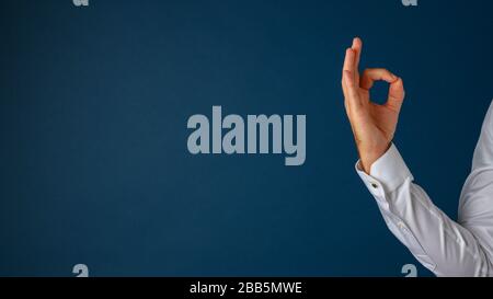 Hand eines Geschäftsmannes, der mit den Fingern eine OK-Geste zeigt. Über blauem Hintergrund mit viel Kopierbereich auf der linken Seite eines Bildes. Stockfoto