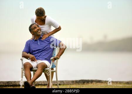 Reifer Mann, der von seiner Frau am Strand eine Schultermassage erhält. Stockfoto