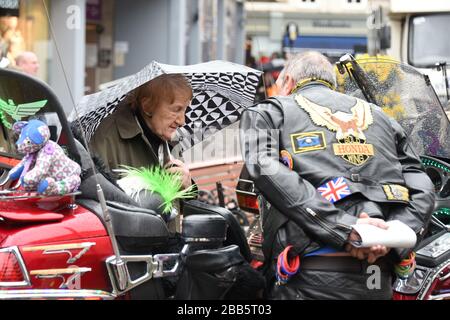 Ältere Frau, die mit einem männlichen Biker spricht, während sie sich ein Motorrad von Honda Goldwing ansieht, das im Regen auf dem Shrewsbury's Town Square geparkt wurde. Stockfoto