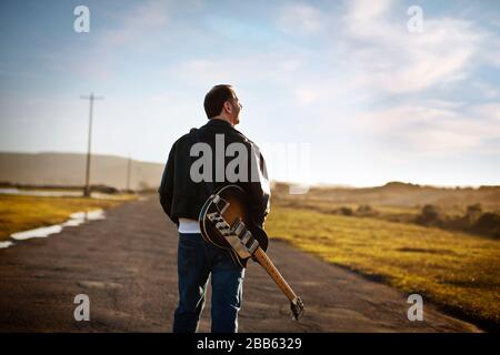 Mann, der auf einer Landstraße mit einer E-Gitarre auf dem Rücken spazieren geht. Stockfoto