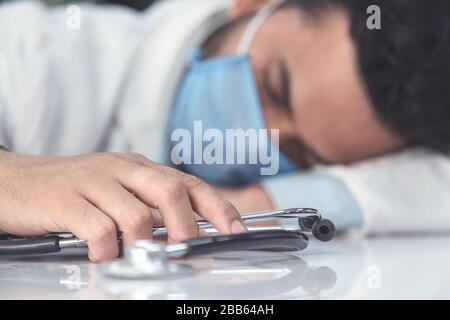 Coronavirus COVID-19 Pandemie. Der müde, erschöpfte Arzt in Uniform schläft nach harter Arbeit im Krankenhausdesk oder in der Klinik, männlich mit Maske und Stethosco Stockfoto